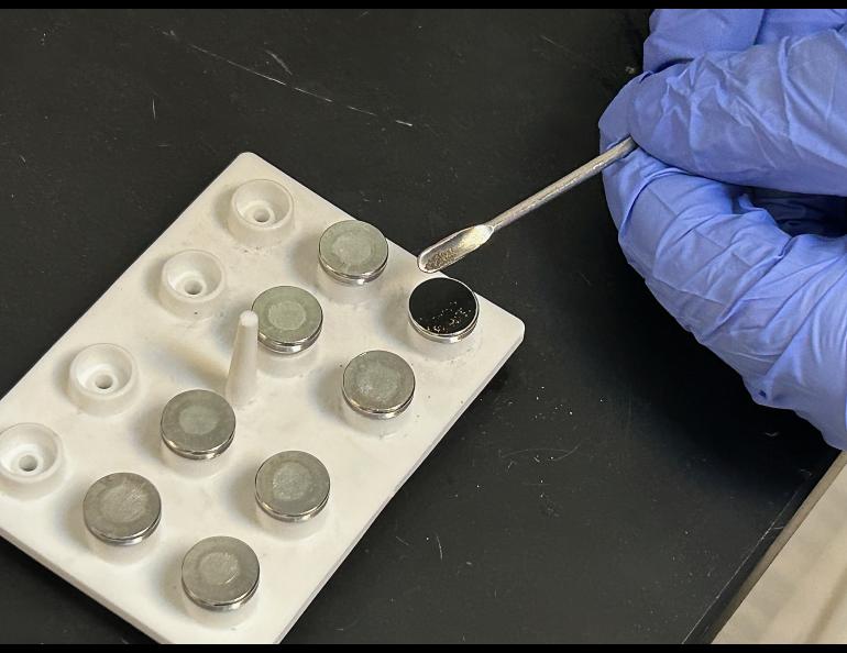 A gloved hand places ash in a scientific instrument to analyze the contents.