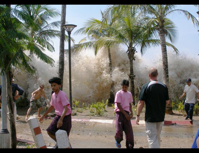 A large wave hits palm trees with people running in the foreground