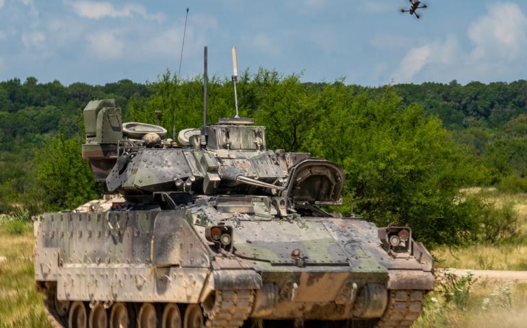 A small quadcopter unmanned aircraft system hovers above an M2 Bradley Infantry Fighting Vehicle on a dirt training range at Fort Hood, Texas, with trees in the background. The scene, captured Aug. 27, 2025, during Operation Return of the Condor, shows the drone positioned overhead in a test.