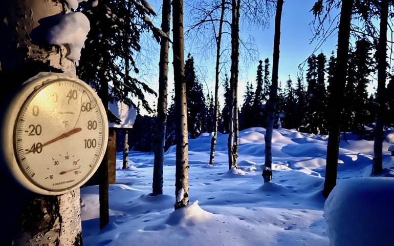 A round outdoor thermometer mounted on a snow-covered tree reads about minus 45 degrees Fahrenheit in a quiet, snow-blanketed forest of tall trees. Long blue shadows stretch across the deep snow under a clear sky. The image shows extreme cold conditions at a home in Two Rivers, Alaska.