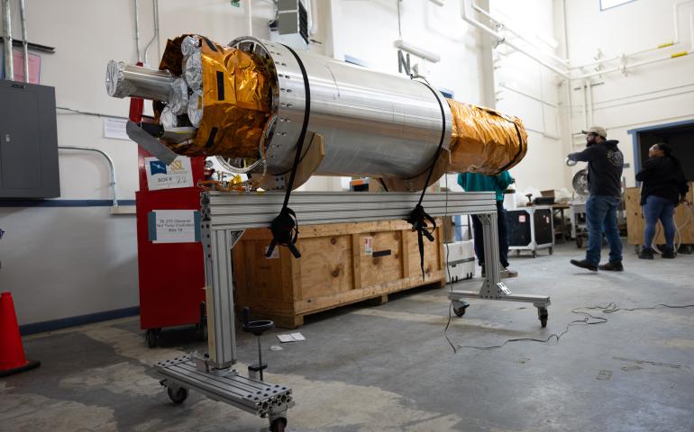 The payload section of the FOXSI-5 mission rests on a wheeled support frame inside the payload assembly building at Poker Flat Research Range. The cylindrical instrument, wrapped partly in gold thermal insulation, is surrounded by equipment crates and technicians preparing it for flight. 