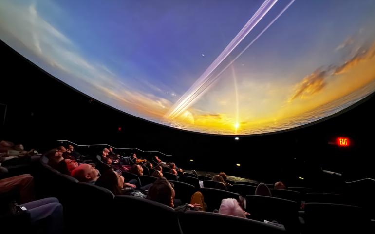 Audience members sit in reclined theater seats, looking up at a curved dome screen displaying a vivid space scene during a movie screening. The image shows the interior of the new planetarium, where attendees enjoy a pre-opening event March 31, with the immersive image filling the entire ceiling.
