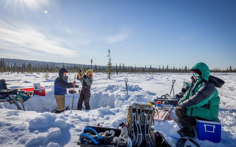 Three bundled researchers work at a snow-covered permafrost coring site in a boreal forest near Fairbanks under a bright, low winter sun. They are operating coring equipment beside a dug trench, with shovels, packs and field gear spread across the snow in the Goldstream Creek area on April 13, 2026.
