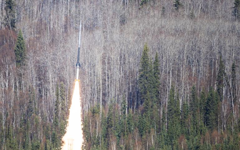 A NASA sounding rocket carrying the FOXSI-5 mission lifts off from Poker Flat Research Range north of Fairbanks at 11:23 a.m. Thursday, May 14, 2026. A bright plume of flame and smoke rises from the launch pad as the rocket climbs above a dense forest of spruce and leafless birch trees.