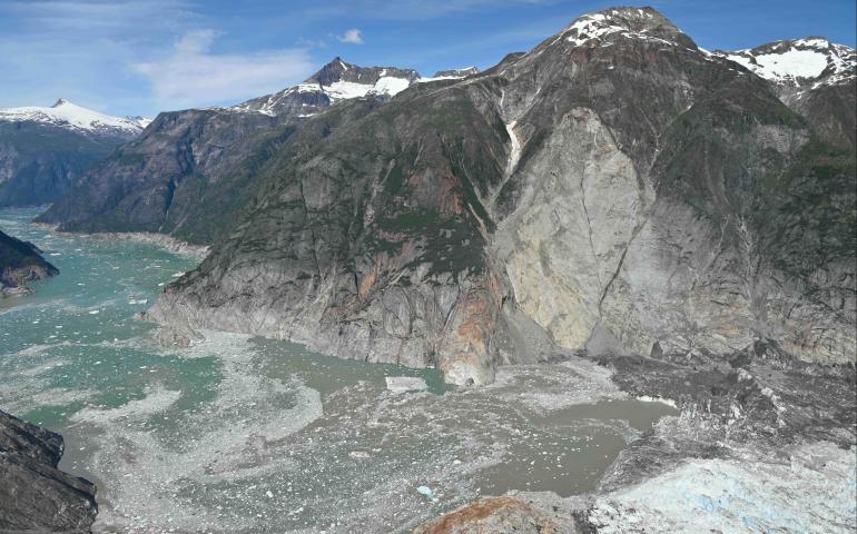 A wide aerial view shows the landslide scar, descending from a steep mountainside into the fjord near a glacier terminus. Gray rock and debris cut through the center of the mountain, reaching the water below, where floating ice and sediment spread across the fjord.