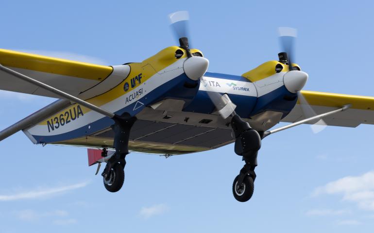 ACUASI’s yellow, white and blue Windracers ULTRA drone carrying chilled human blood samples takes off from Nenana Municipal Airport on Aug. 19, 2026. The twin-engine aircraft climbs with its landing gear still extended against a bright blue sky with thin clouds.