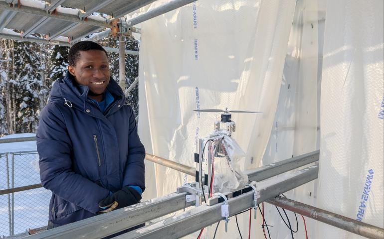 Doctoral student Morakinyo Labiran stands at ACUASI’s ice tower beside his planned test item, a small rotor assembly mounted on metal rails and surrounded by wiring. He wears a heavy blue winter coat and gloves inside the translucent plastic-lined, scaffold-supported enclosure.