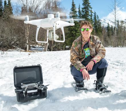 Jen Schmidt, woman with short light brown hair, crouches in the snow next to a drone control unit. A white drone hovers in the air between her and the camera.