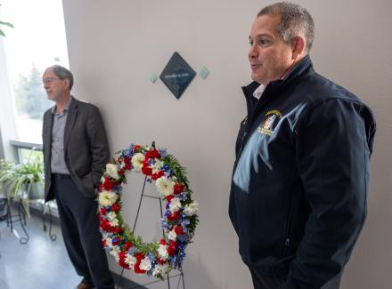 Two men stand on either side of a wreath with red, white and blue flowers on a stand in the Elvey hall.