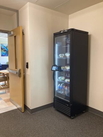 A tall vending machine with refrigerated food items stands in a corner. 