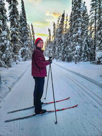 A man on cross-country skis on a path through the forest looks back at the camera and smiles.