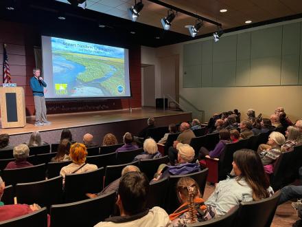 Ken Tape, research professor at the Geophysical Institute, speaks from a stage to a packed house at the Morris Thompson Cultural & Visitors Center.