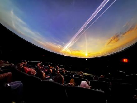A round movie screen shows the sky as an audience sits in the theater below it