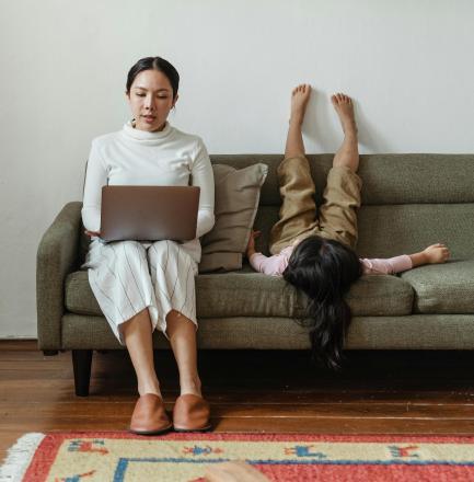 Woman sitting on couch with computer on her lap and a toddler next to her, feet up on the wall