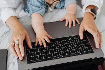 Hands of an adult in front of a laptop computer with a baby in their lap who also has a hand on the keyboard