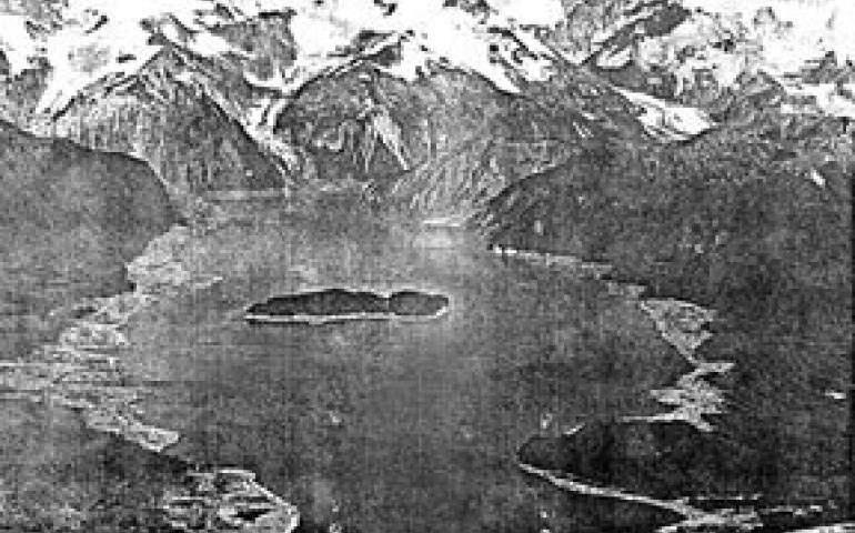 The central portion of T-shaped Lituya Bay with Cenotaph Island in its center. Mt. Crillion, 12, 726 feet high, towers over other peaks surrounding the bay. Treeless areas of the shore were wiped clean by the wave which swept down the bay on July 10, 1958. Photo by Byron Hale.