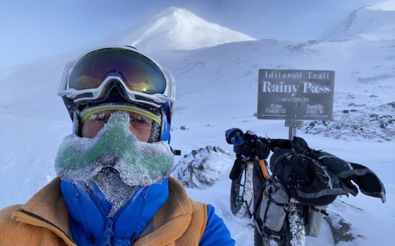 Peter Delamere poses in front of a sign in Rainy Pass in the Alaska Range during the 2024 Iditarod Trail Invitational race. On his face is a “nose-hat” invented by Fairbanks athlete Shalane Frost. Photo by Peter Delamere.