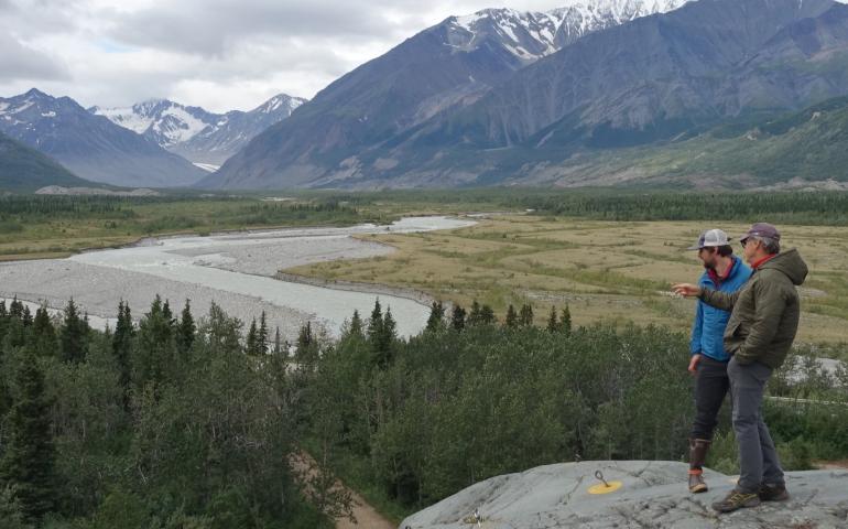 Phillip Wilson (blue jacket) and Dan Mann stand on a rock outcrop that was scoured by floodwaters a few centuries ago when Black Rapids Glacier — far in the distance — advanced to dam the Delta River. Photo by Ned Rozell.