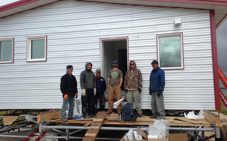 Aaron Cooke, in the T-shirt and blue hat, poses in front of an efficient house he helped design after he and Newtok residents built it at the new village site of Mertarvik in 2016. Photo by Molly Rettig. 