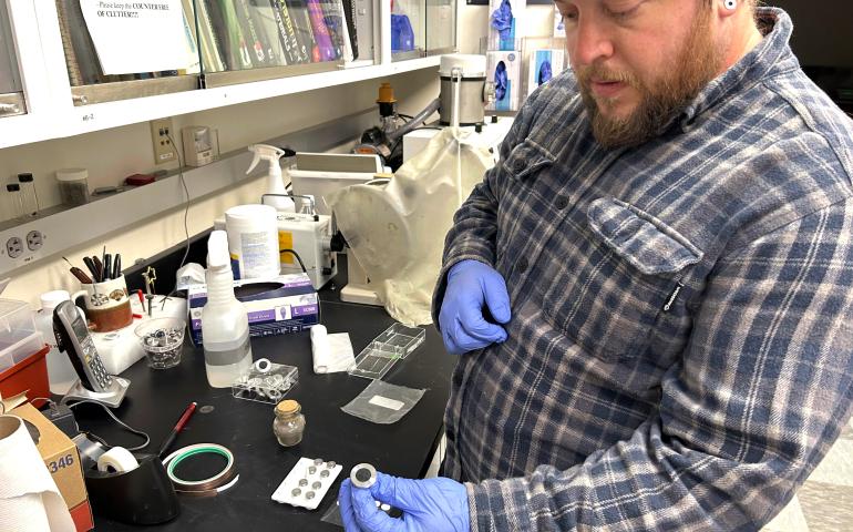 Nathan Graham, director of the UAF Geophysical Institute’s Advanced Instrumentation Lab, holds a mounted sample of Mount Spurr ash in a laboratory filled with sample containers, tools and other equipment. The ash will be analyzed with a scanning electron microscope.