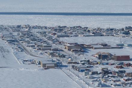 Aerial view of Utqiaġvik, Alaska, showing a snow-covered coastal town bordered by sea ice. A long, narrow strip of open water called an open flaw lead cuts across the frozen ocean, separating the solid landfast ice attached to shore from the drifting pack ice offshore. Photo by Andrew Mahoney