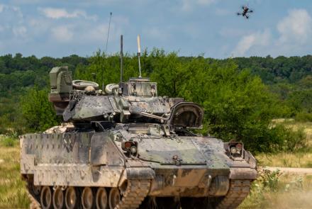 A small quadcopter unmanned aircraft system hovers above an M2 Bradley Infantry Fighting Vehicle on a dirt training range at Fort Hood, Texas, with trees in the background. The scene, captured Aug. 27, 2025, during Operation Return of the Condor, shows the drone positioned overhead in a test.