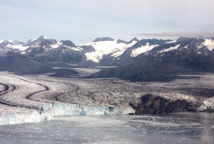 A wide view of Columbia Glacier in Alaska shows a broad river of ice flowing between dark, rocky mountains, with snow-covered peaks in the distance and floating ice in the water at the glacier’s front. 