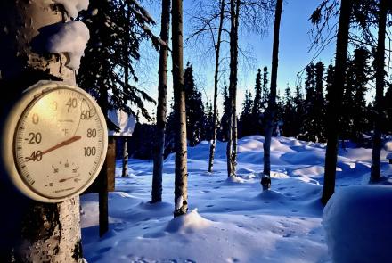 A round outdoor thermometer mounted on a snow-covered tree reads about minus 45 degrees Fahrenheit in a quiet, snow-blanketed forest of tall trees. Long blue shadows stretch across the deep snow under a clear sky. The image shows extreme cold conditions at a home in Two Rivers, Alaska.