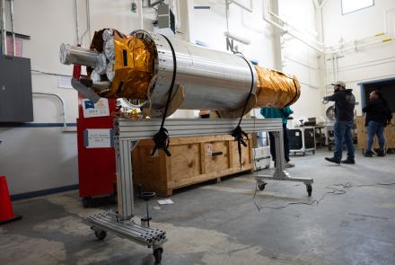 The payload section of the FOXSI-5 mission rests on a wheeled support frame inside the payload assembly building at Poker Flat Research Range. The cylindrical instrument, wrapped partly in gold thermal insulation, is surrounded by equipment crates and technicians preparing it for flight. 