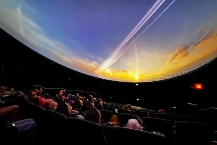Audience members sit in reclined theater seats, looking up at a curved dome screen displaying a vivid space scene during a movie screening. The image shows the interior of the new planetarium, where attendees enjoy a pre-opening event March 31, with the immersive image filling the entire ceiling.