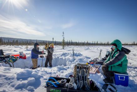 Three bundled researchers work at a snow-covered permafrost coring site in a boreal forest near Fairbanks under a bright, low winter sun. They are operating coring equipment beside a dug trench, with shovels, packs and field gear spread across the snow in the Goldstream Creek area on April 13, 2026.
