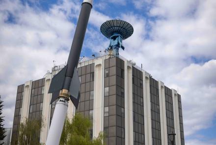 Visitors on the Geophysical Institute summer tour will learn about the large blue satellite antenna atop the Elvey Building and the sounding rocket installation displayed on the lawn outside. The photo shows the rocket model rising in the foreground in front of the building under a partly cloudy sky