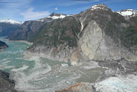 A wide aerial view shows the landslide scar, descending from a steep mountainside into the fjord near a glacier terminus. Gray rock and debris cut through the center of the mountain, reaching the water below, where floating ice and sediment spread across the fjord.