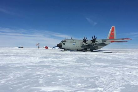 A New York Air National Guard LC-130 Skibird cargo aircraft sits on a snow runway atop the Greenland Ice Cap under a clear blue sky. The ski-equipped aircraft, operated by the 109th Airlift Wing, is parked on the windswept ice with a small field camp, flags and support structures visible.