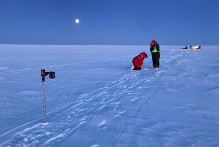  Two researchers in heavy winter clothing kneel and stand on the snow-covered surface of Teshekpuk Lake as they conduct ice measurements under a moonlit Arctic sky. An ice auger stands in the foreground near snowmachine tracks, while additional team members and equipment are in the distance. 