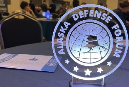 A glowing Alaska Defense Forum sign sits on a table at the 2025 event at the 8 Star Event Center in Fairbanks. The circular sign features an outline of Alaska over a globe, with conference attendees visible in the background.