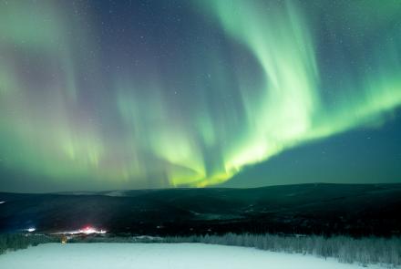 Green aurora curtains stretch across the night sky above snow-covered hills at Poker Flat Research Range north of Fairbanks in late January 2025. Facility lights glow in the valley below.