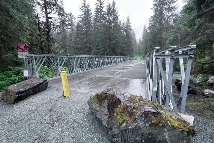 Rainy silver metal pedestrian bridge crosses Montana Creek in Juneau, surrounded by dense evergreen forest. The wet metal bridge deck leads into the trees, with the creek visible below on the right.