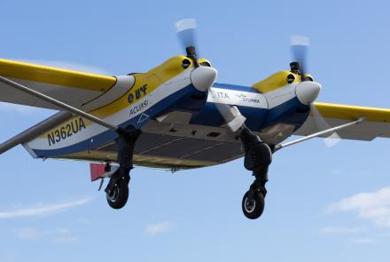 ACUASI’s yellow, white and blue Windracers ULTRA drone carrying chilled human blood samples takes off from Nenana Municipal Airport on Aug. 19, 2026. The twin-engine aircraft climbs with its landing gear still extended against a bright blue sky with thin clouds.
