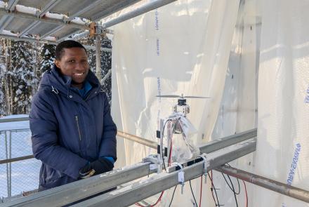 Doctoral student Morakinyo Labiran stands at ACUASI’s ice tower beside his planned test item, a small rotor assembly mounted on metal rails and surrounded by wiring. He wears a heavy blue winter coat and gloves inside the translucent plastic-lined, scaffold-supported enclosure.