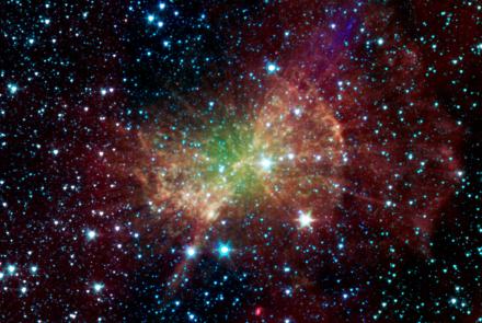 The Dumbbell Nebula glows in infrared light in this 2011 image from NASA’s Spitzer Space Telescope. A broad, irregular cloud of green, yellow and orange gas fills the center, surrounded by wispy red clouds against the blackness of space. Hundreds of bright blue-white stars are scattered throughout.