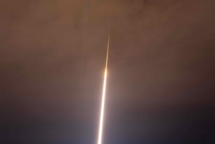 A two-stage NASA Black Brant IX sounding rocket creates a brilliant white exhaust trail that rises from the brightly illuminated launch site above snow-covered hills and forest, extending into orange-tinted clouds, where the rocket appears as a small bright streak.