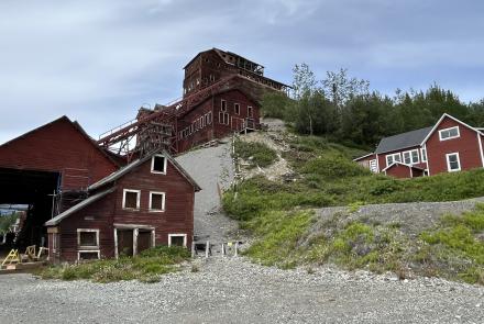 Historic red wooden buildings of the Kennecott Mines complex climb a green mountainside in Wrangell-St. Elias National Park and Preserve. The towering copper mill processed ore from the Bonanza, Jumbo, Mother Lode, Erie and Glacier mines; the restored mine office stands at right.