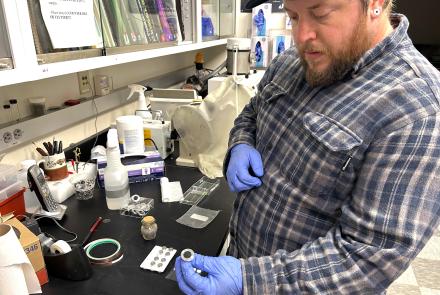 Nathan Graham, director of the UAF Geophysical Institute’s Advanced Instrumentation Lab, holds a mounted sample of Mount Spurr ash in a laboratory filled with sample containers, tools and other equipment. The ash will be analyzed with a scanning electron microscope.