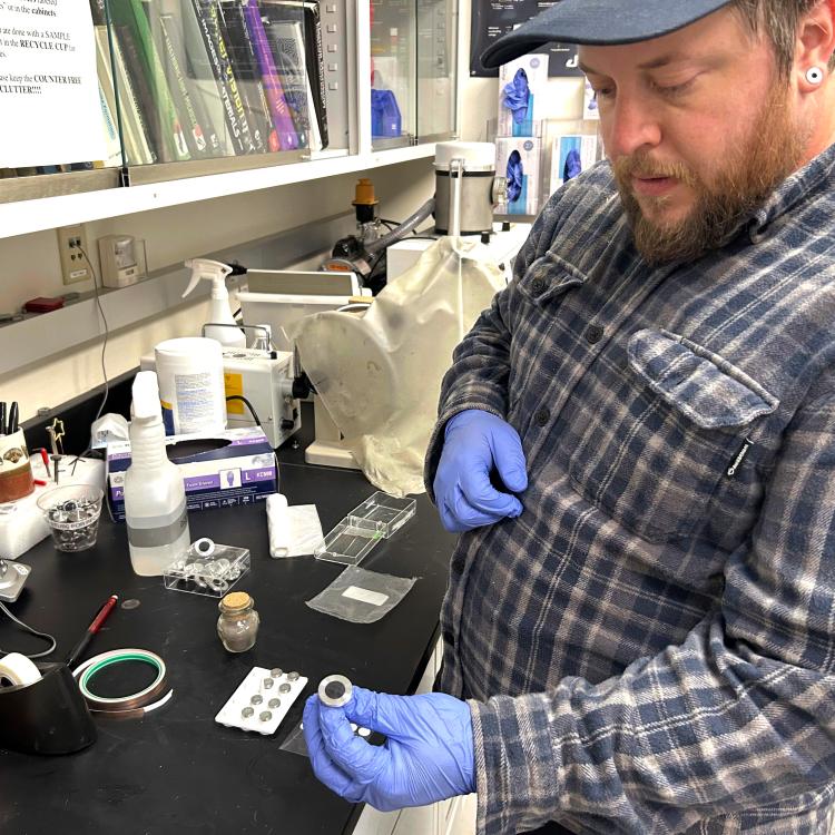 Nathan Graham, director of the UAF Geophysical Institute’s Advanced Instrumentation Lab, holds a mounted sample of Mount Spurr ash in a laboratory filled with sample containers, tools and other equipment. The ash will be analyzed with a scanning electron microscope.